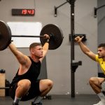 Athlete performing weightlifting exercise under trainer's guidance in a gym setting.